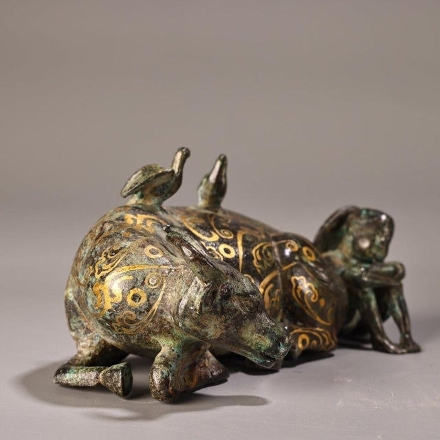 This is a cast bronze figurine featuring a reclining ox accompanied by a small human figure (a herder) seated at its rear....