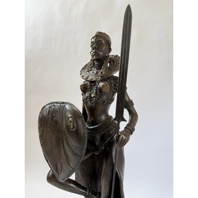 This bronze sculpture presents a powerful African female warrior rendered in a poised yet commanding stance, holding a...