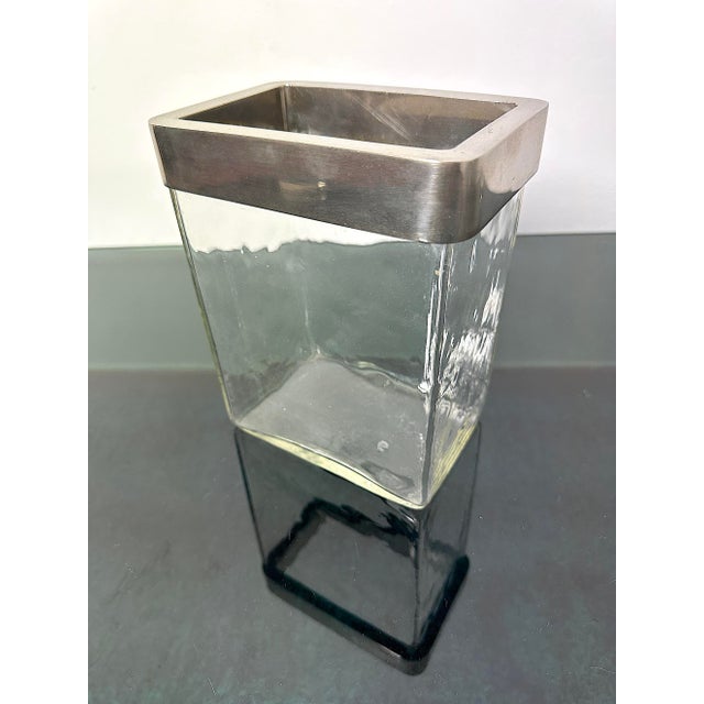 Flower vase composed of a transparent glass container with a rectangular base and a decorative upper edge in satin metal....
