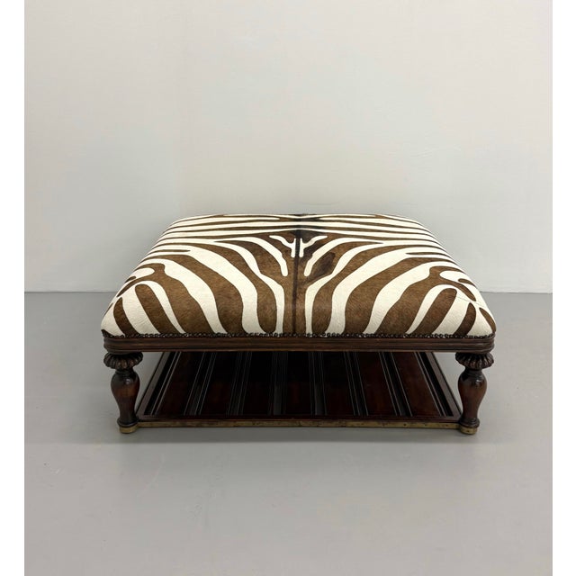 Statement ottoman featuring zebra hide upholstery, classic nailhead trim, and a dark wood frame with turned legs and...