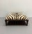 Statement ottoman featuring zebra hide upholstery, classic nailhead trim, and a dark wood frame with turned legs and...