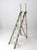 An antique wooden ladder with a folding form, crafted with sturdy wood framing and side rails featuring open window...