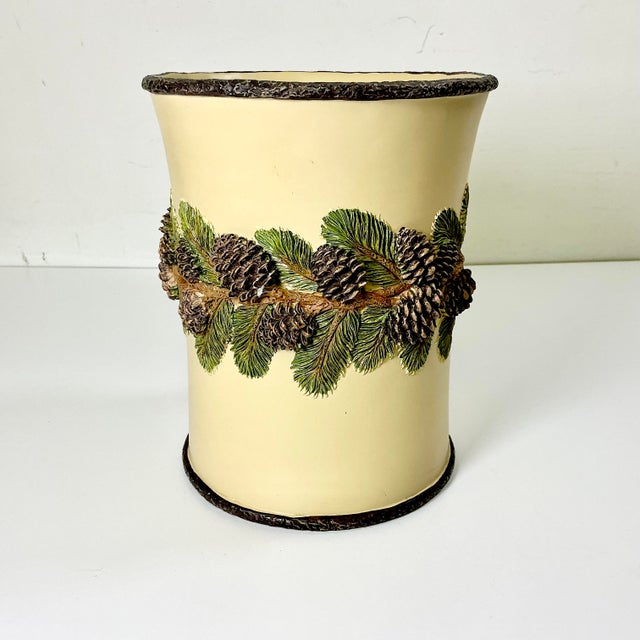 This is an attractive Rustic wastebasket. It has a ring of dark green pine tree branches with brown pinecones encircling...