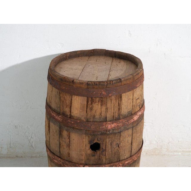 This antique wooden barrel with iron hoops features a cylindrical body of oak staves held by hand-forged iron hoops and a...