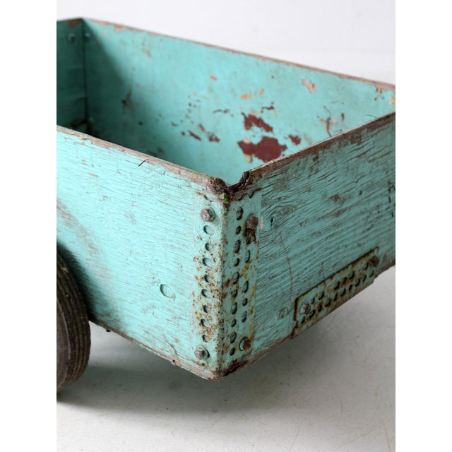 This vintage wooden garden cart features a worn teal finish, two side wheels, and a metal handle for easy maneuvering....