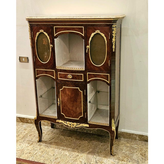 This elegant Louis XV-style display cabinet is a refined statement piece crafted in richly toned wood with exquisite...