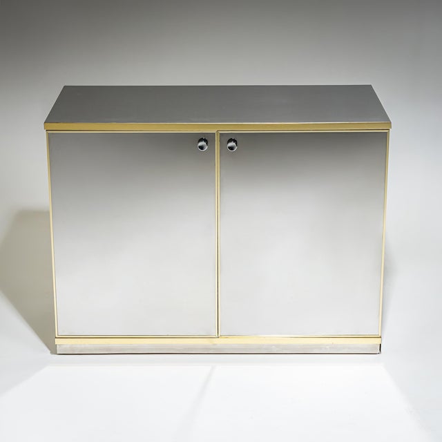 Pair of two-door sideboards in the Hollywood Regency style, featuring mirrored surfaces and a contrasting brass trim. The...