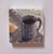 This oil on board painting captures a simple still life featuring a dark mug alongside a slice of citrus. The loose...