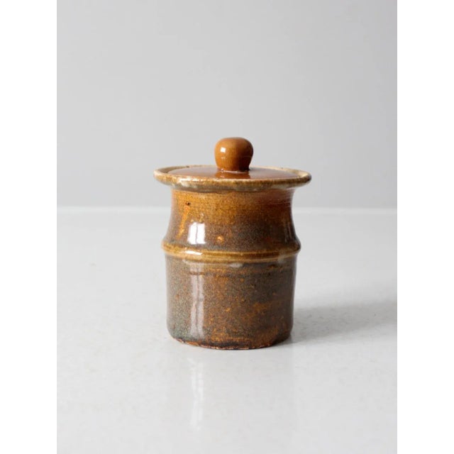 This vintage studio pottery jar is a small, handmade piece featuring a rich, variegated earthy brown glaze. The lid has a...