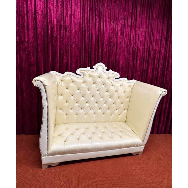 Statement loveseat featuring ivory button-tufted upholstery, an ornate white frame, a decorative crest, and a dramatic...