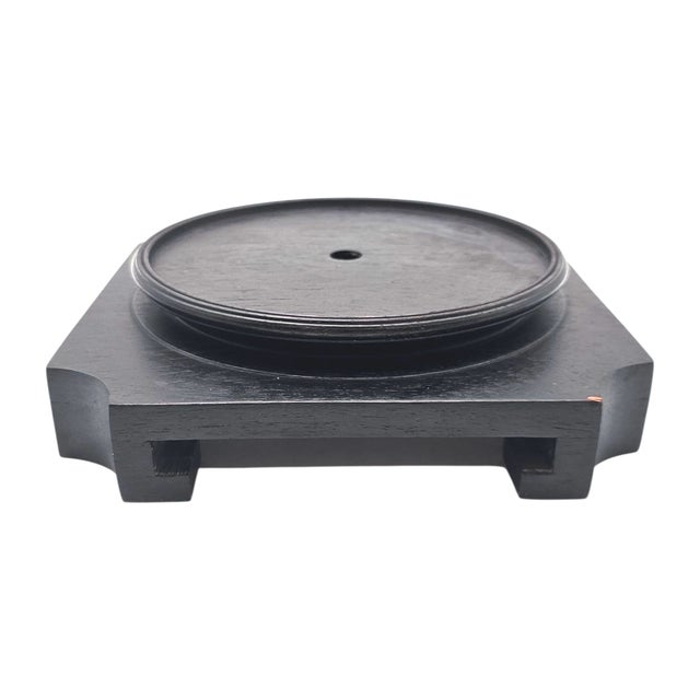 A square base of ebony, dark and steady, carved with restraint to hold vessel or sculpture. The grain runs deep, polished...
