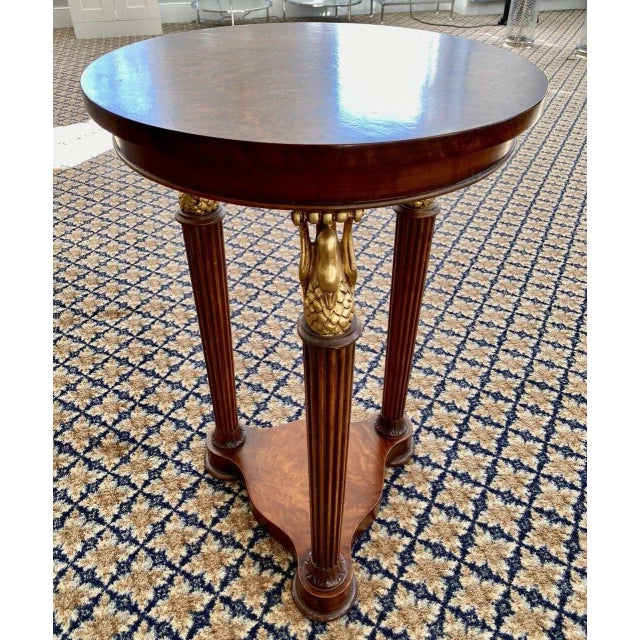 An elegant neoclassical mid-century pedestal occasional side table with a beautiful burl wood top sitting above a trio of...
