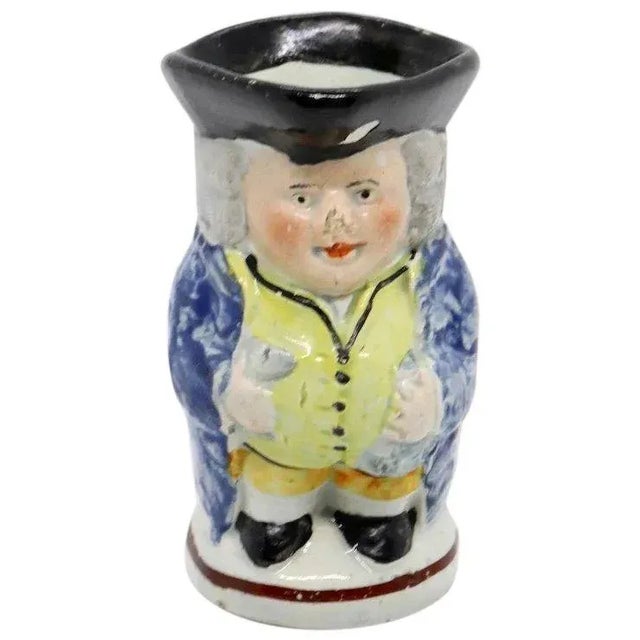 DETAILS An antique Toby Jug of traditional form with a standing man, wearing a blue coat, yellow vest, breeches and a...