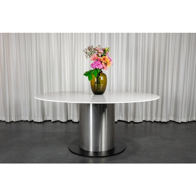 Large round dining room table composed of a white Corian top resting on a central stainless steel post. This table is in...