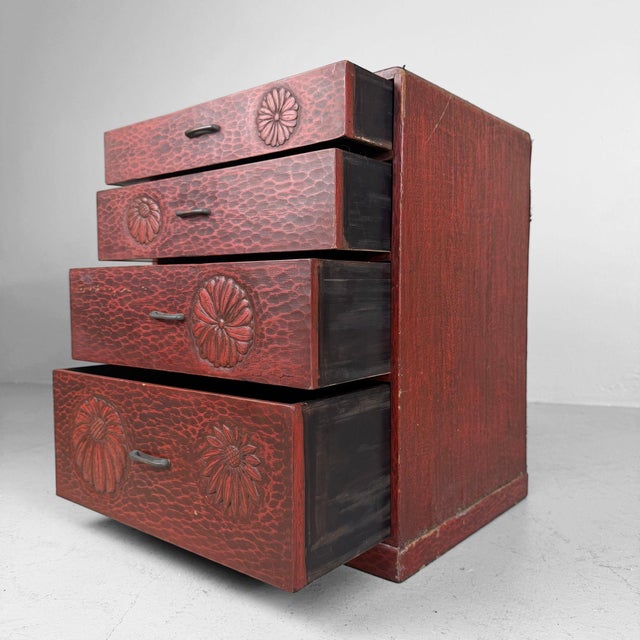 A compact Japanese drawer cabinet with four drawers, a deep reddish-brown colour and decorative floral motifs in relief....