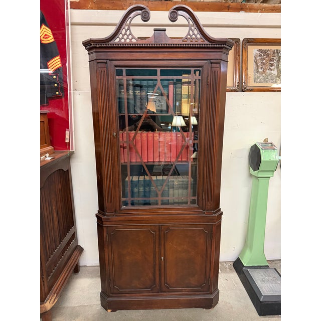 Elegant mahogany corner display cabinet featuring an ornate broken-scroll pediment, geometric astragal glass door, and...