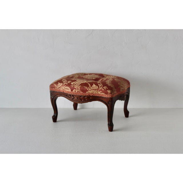 Vintage French Louis XV-style carved wooden footstool upholstered in a rich red and gold floral fabric. The cushion is...