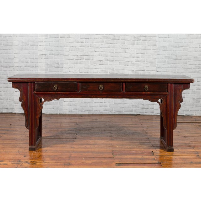 A Chinese Qing Dynasty period elmwood low console table from the 19th century, with three drawers and carved spandrels....
