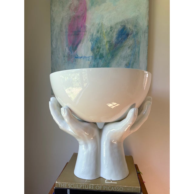 Striking Italian ceramic centerpiece featuring a large white bowl cradled by a sculptural pair of hands. The glossy white...