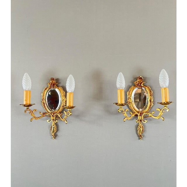 An elegant pair of gilt bronze wall sconces, each adorned with an oval beveled mirror. The two candle-shaped arms evoke...