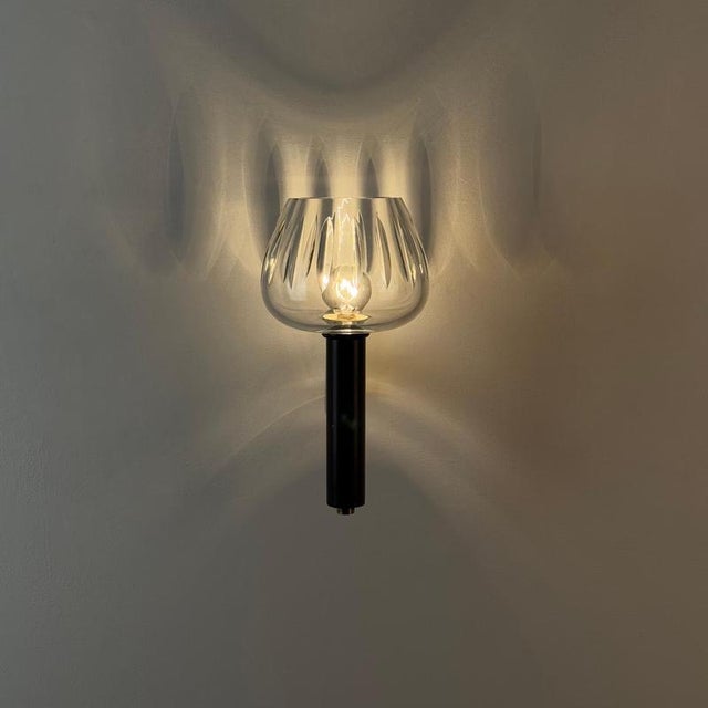 A quietly theatrical wall light, defined by the tension between strict geometry and a soft, refracted glow. The silhouette...