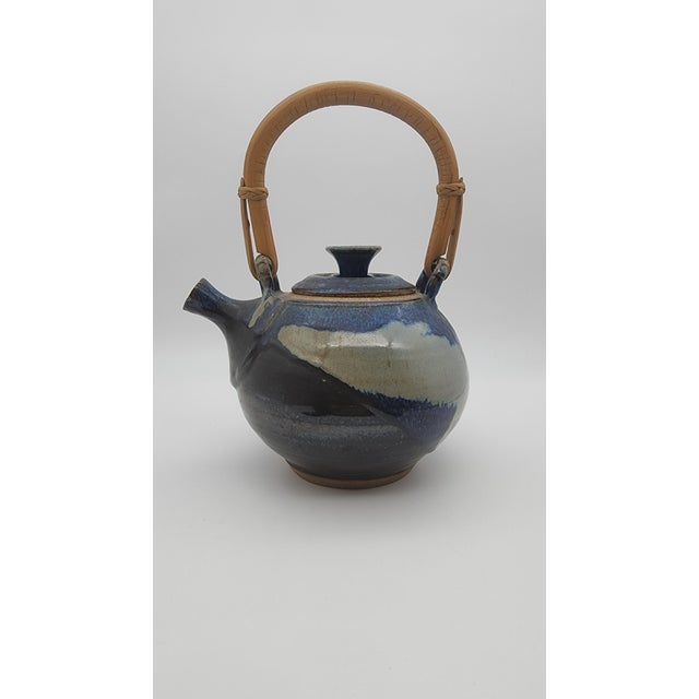 A sculptural studio pottery teapot featuring a rounded body, expressive cobalt and cream drip glaze, and a woven bentwood...