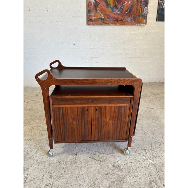 Gorgeous mid century modern rolling bar cart, featuring sculpted rosewood with a black laminate inlay top that extends...
