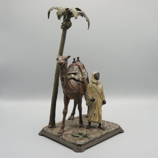 A cold painted and patinated bronze sculpture depicting a bedouin man astride his camel standing under a palm tree....