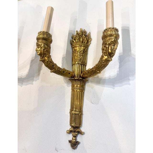 Pair of French Empire gilt bronze two-light sconces, 19th century, with a flame atop and empire faces design