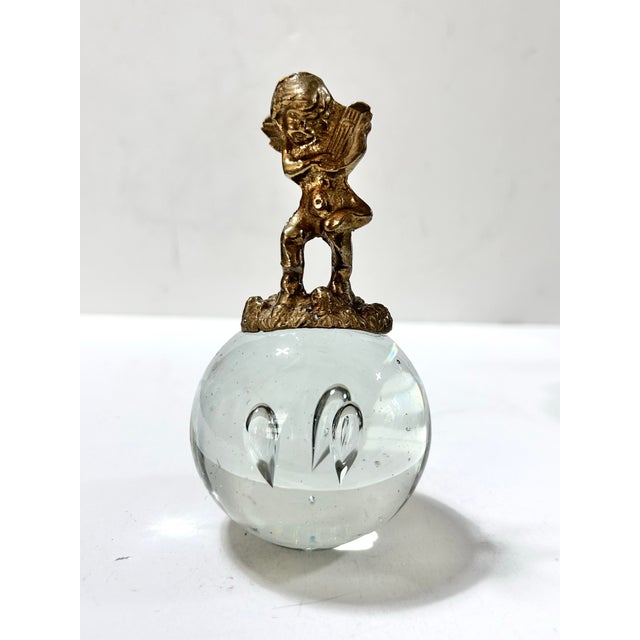 A gold-tone cherub playing a lyre sits atop a clear glass orb with three suspended teardrop-shaped bubbles. A decorative...