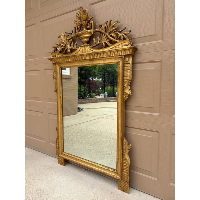 An elegant Italian giltwood mirror featuring a carved urn crest, foliate and rope swags, and Greeg key details. The...