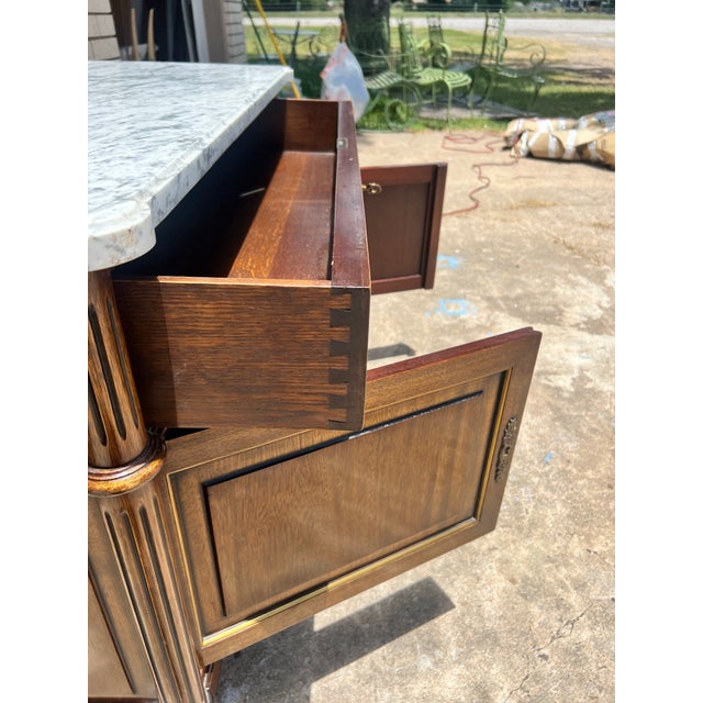 An elegant Louis XVI style commode featuring a warm wood finish with a white marble top. The piece showcases clean lines,...