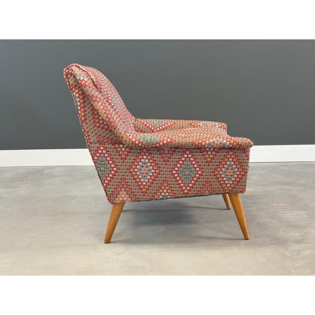 This mid century modern lounge chair features a bold, woven fabric with a geometric diamond pattern in warm tones of red,...