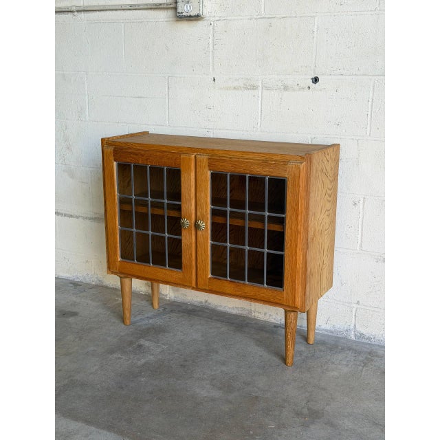 This compact oak cabinet presents a clean rectangular case on four tapered legs. Two leaded glass doors create a...