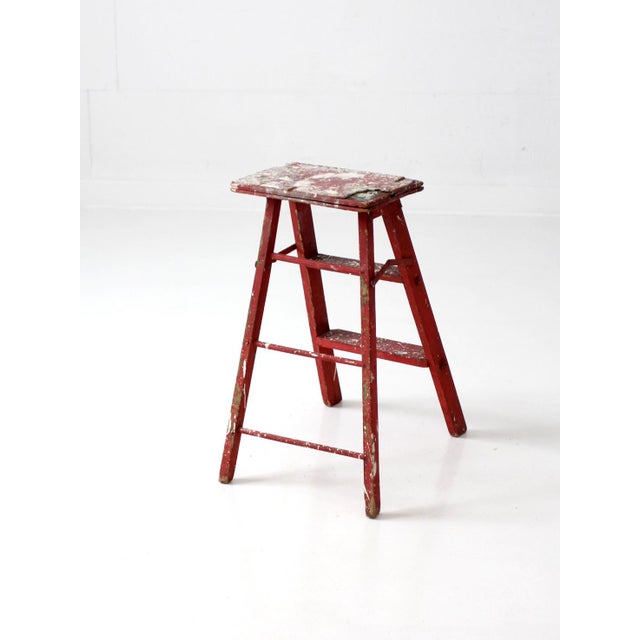 Vintage wooden step ladder featuring a weathered red painted finish, authentic paint splatter, and a beautifully aged...
