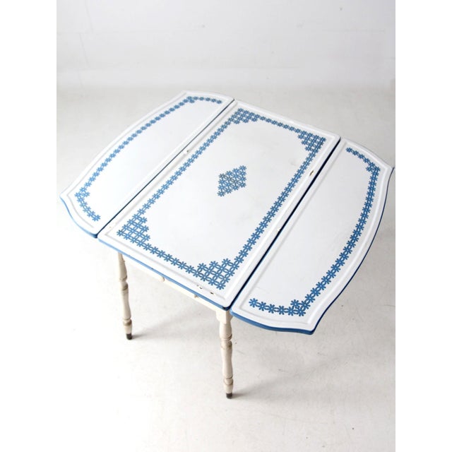 This 1930s–1940s antique drop-leaf table features a white enamel top bordered in blue with a delicate floral pattern. The...