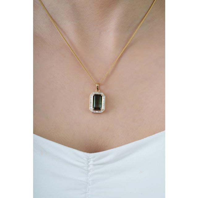 This halo pendant showcases a striking 5.5 carat emerald-cut green tourmaline, set in 14K yellow gold. The tourmaline...