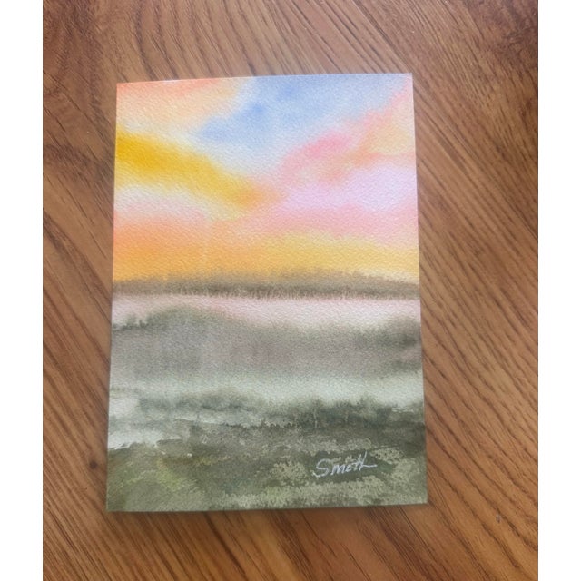 Original watercolor capturing a glowing sunset sky over a quiet, open landscape. Warm yellows, peach, and soft blue tones...