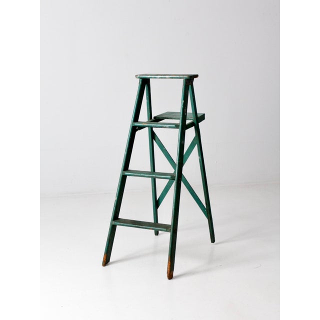 This is a vintage green wooden ladder. The painted a-frame ladder features three grooved steps, a back shelf, and crossing...