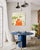 Food Lady depicts a seated female figure dressed in vivid orange, set within a landscape of green hills, wind turbines,...