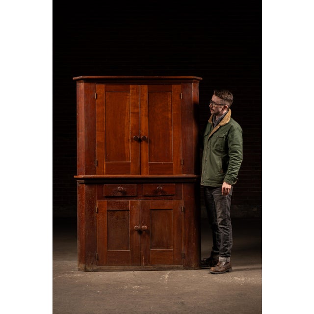 A substantial two-piece primitive corner cabinet constructed in solid wood with a warm reddish-brown finish and simple...