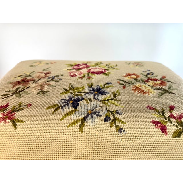 A charming vintage footstool bench featuring hand-stitched floral needlepoint upholstery in a soft, muted palette set...