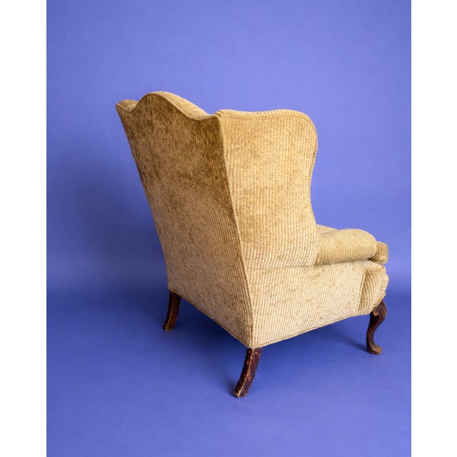 Classic wingback armchair upholstered in a soft, neutral corded fabric. Features rolled arms, a cushioned seat, and carved...