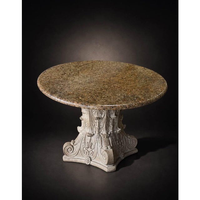 A dramatic round dining table featuring a polished natural stone top and an elaborately detailed pedestal base. The...