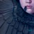 The artwork "Eat" presents a close-up view of a figure adorned in dark, feather-like textures that envelops the face,...