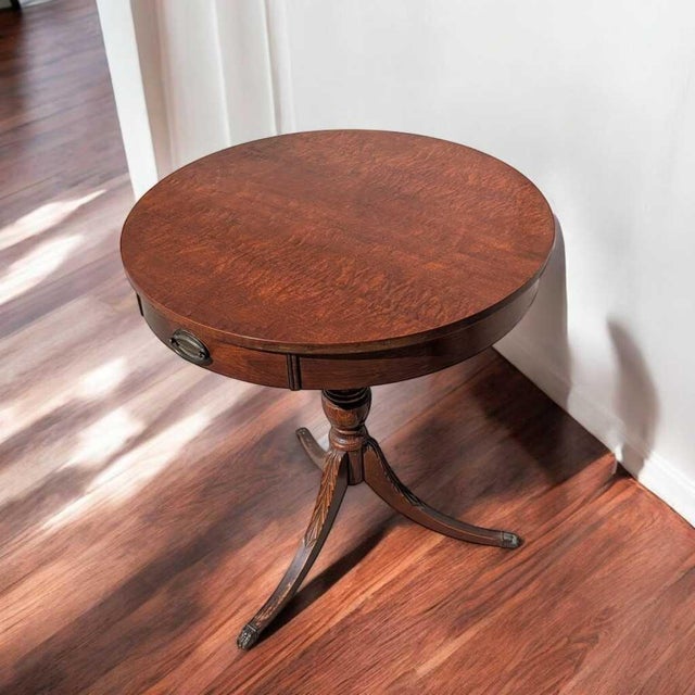 A circular vintage side table by Mersman with a warm wood finish, compact drawer, and a graceful pedestal base on three...