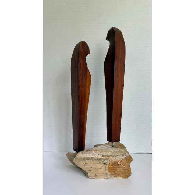 This signed studio sculpture features two abstracted wooden figures mounted on a natural, stepped stone base. Each figure...
