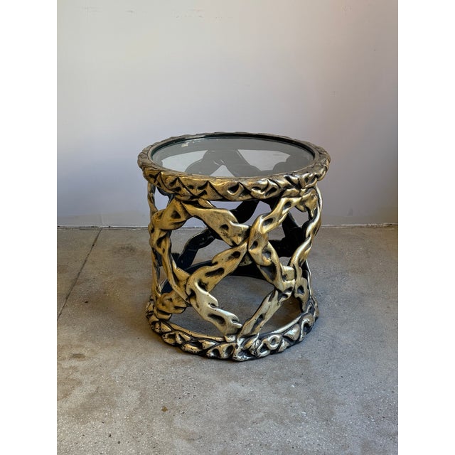 Cast resin side table with an openwork base composed of intertwined, ribbon-like forms, finished in a gilt tone with...