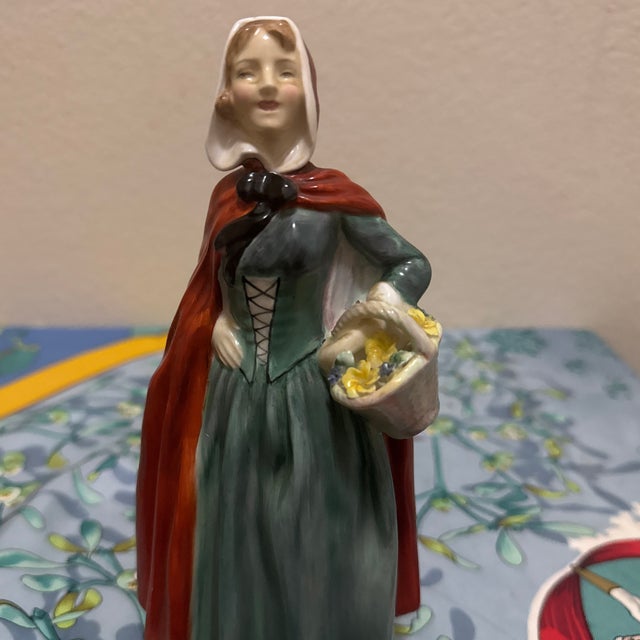 The figurine portrays an elegant lady in a mid-20th-century interpretation of historical attire. She wears a teal or blue-...