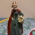 The figurine portrays an elegant lady in a mid-20th-century interpretation of historical attire. She wears a teal or blue-...